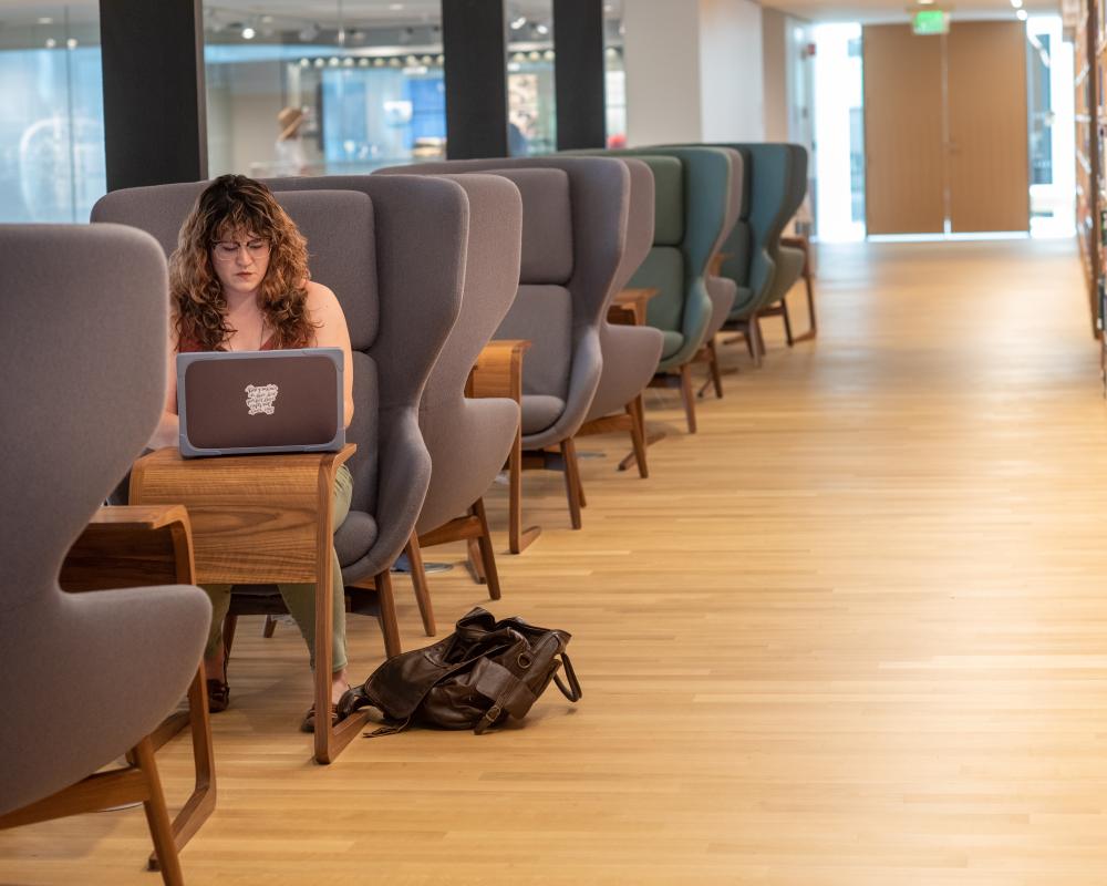 A student sits in a highback chair in the library working on their laptop.